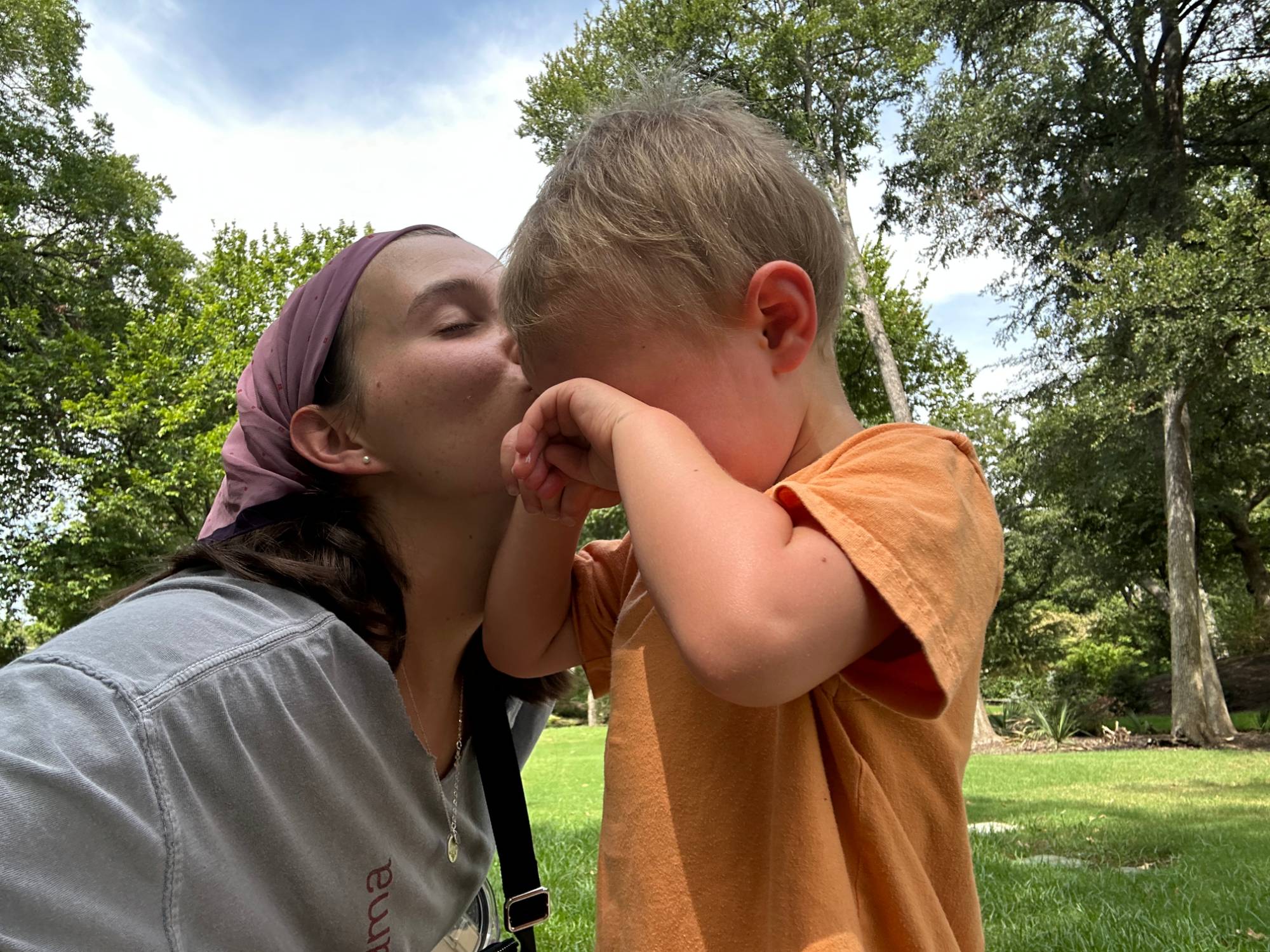 A mother kissing her toddler's cheek in a sunny park